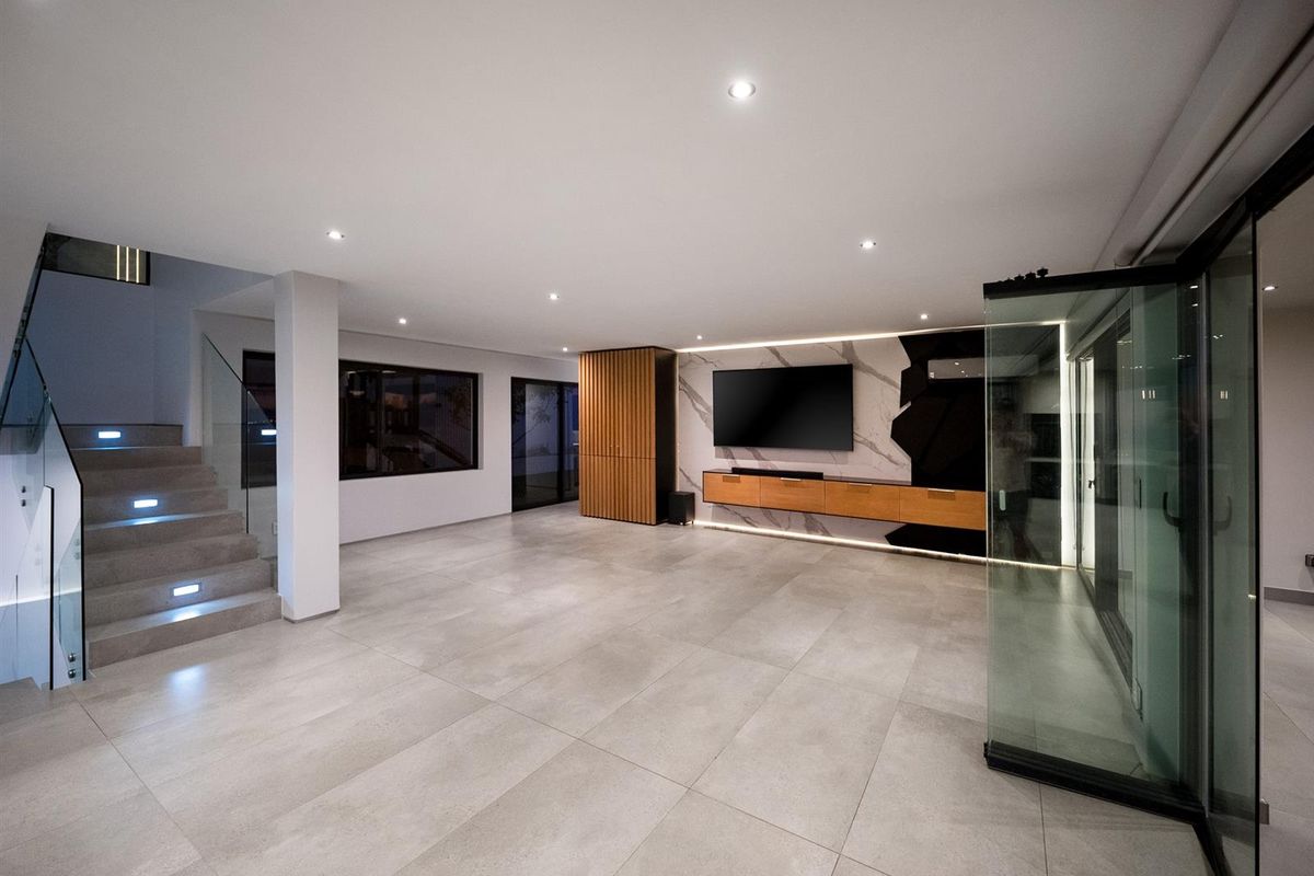 Spacious modern living area featuring large format floor tiles, a built-in media console with marble-effect wall cladding, and a contemporary staircase with glass balustrades.
