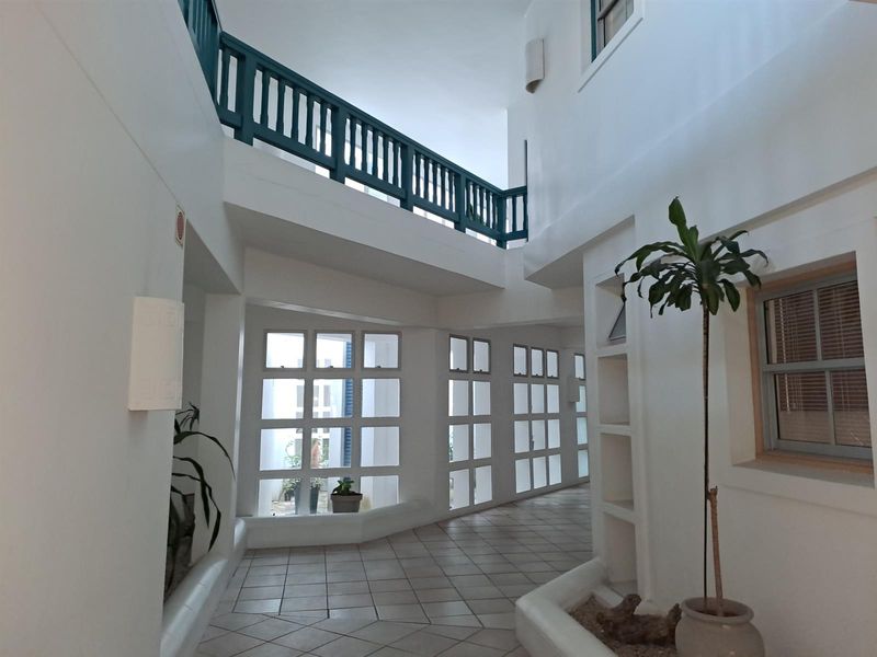 A bright, multi-level interior hallway featuring tiled flooring, built-in planter boxes, and a mezzanine walkway with a decorative teal railing.