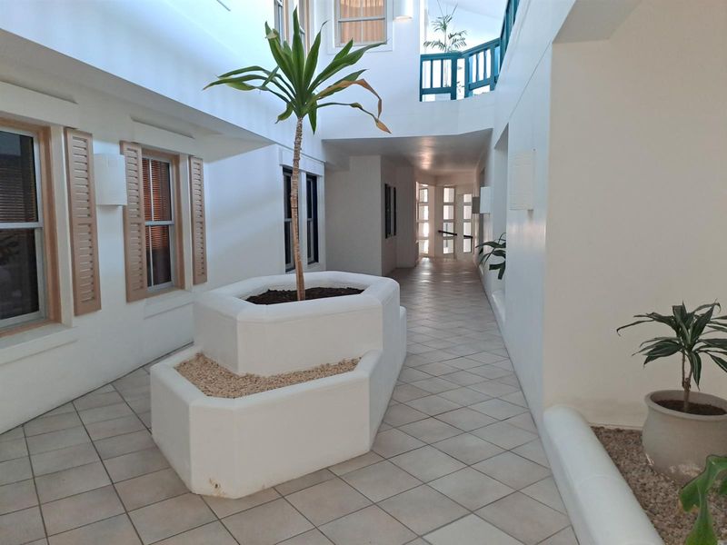 A bright, open-air atrium or hallway featuring tiled flooring, built-in planters, and architectural windows overlooking the central space.