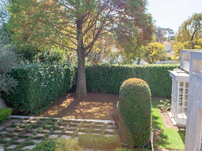 A well-maintained private garden featuring mature trees, manicured hedges, and a paved pathway, viewed from an elevated position.