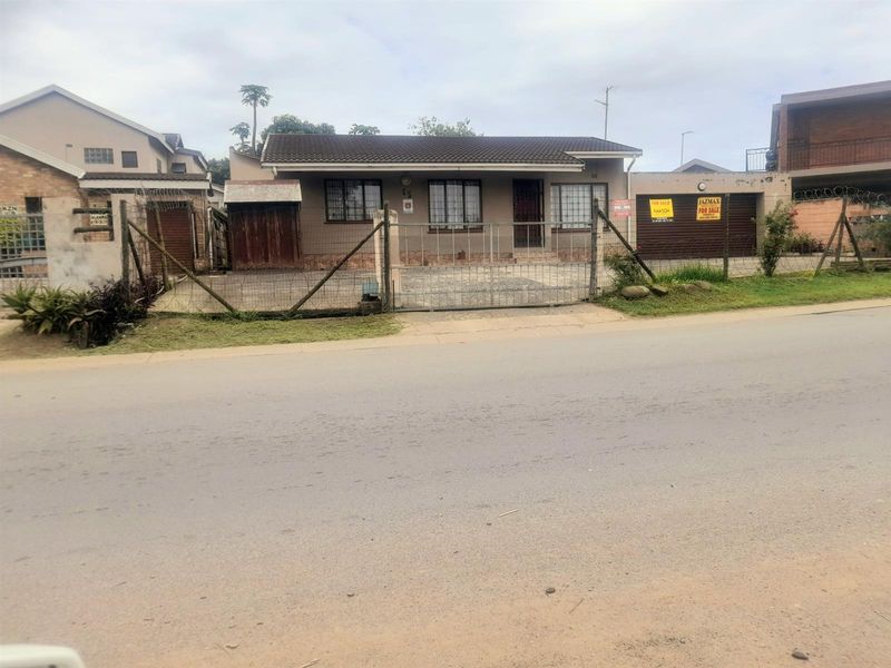 Front elevation of a single-story house featuring a tiled roof, paved driveway, and secure perimeter fencing with a metal gate.