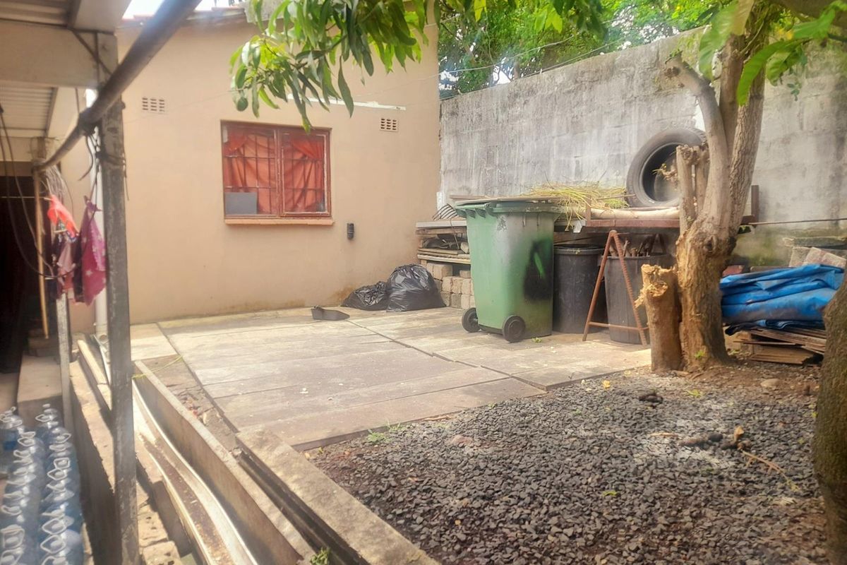 Rear exterior area featuring concrete paving, a boundary wall, and a metal-framed window with security bars, requiring maintenance and clearing.