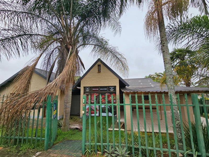 Front view of a residential property featuring a pitched roof, covered patio area, and perimeter security fencing with established palm trees.
