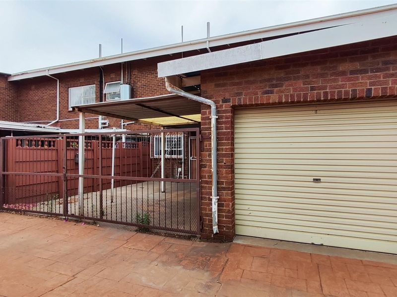 Exterior view of a townhouse featuring a brick facade, a single roll-up garage door, and a paved patio area with metal security fencing.