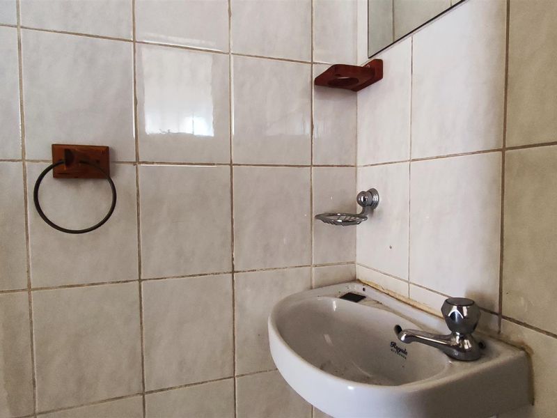 A dated bathroom vanity area featuring white wall tiles, a wall-mounted basin, and fixed chrome accessories.