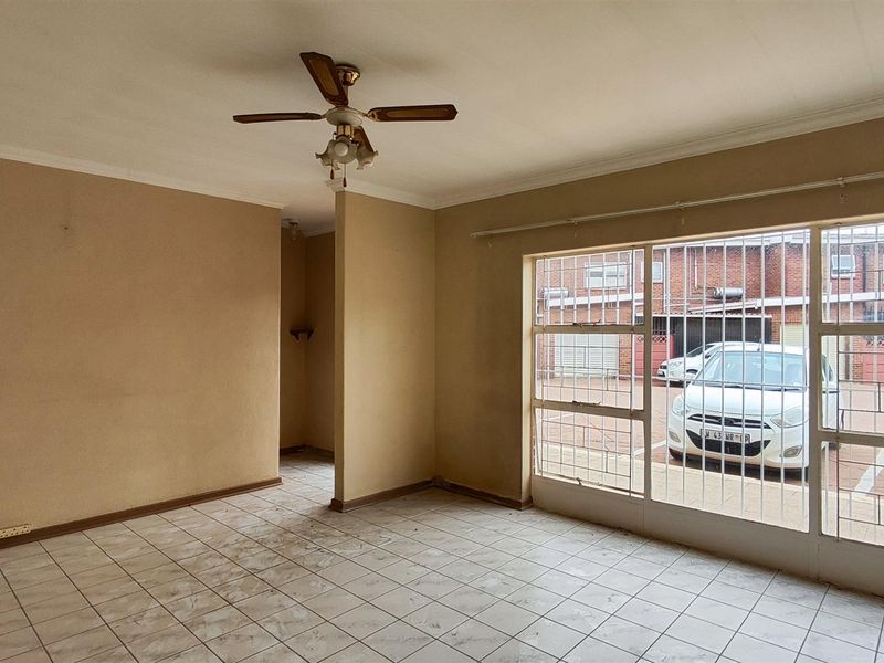 An empty living room featuring tiled flooring, a ceiling fan, and large windows with security bars overlooking the complex parking area.