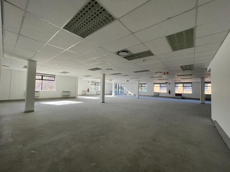 A spacious, open-plan office area featuring a suspended ceiling, recessed lighting, structural columns, and large windows providing ample natural light.