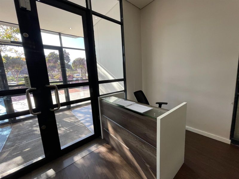 A professional office entrance featuring a modern reception desk, laminate flooring, and large black-framed glass doors providing natural light.