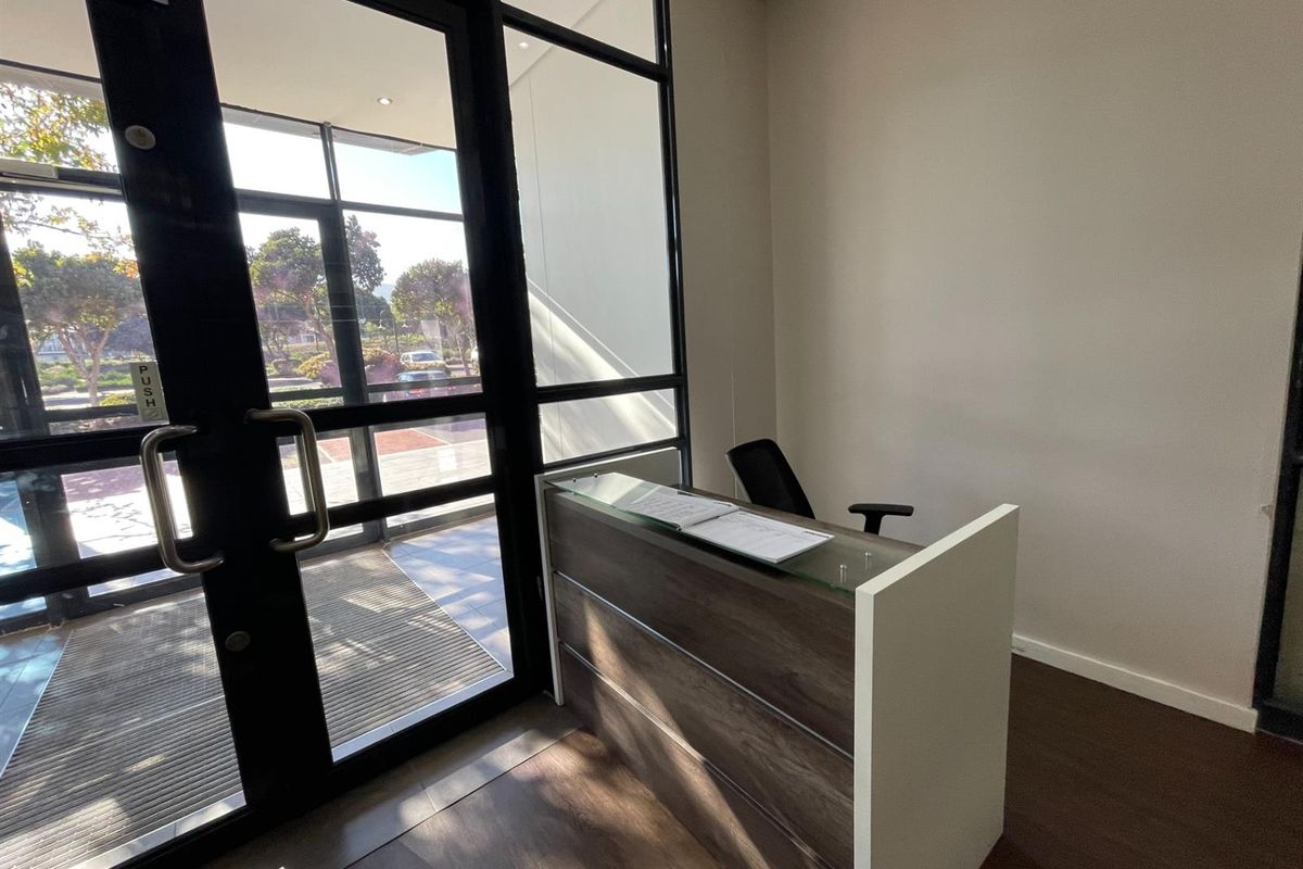 A professional office entrance featuring a modern reception desk, laminate flooring, and large black-framed glass doors providing natural light.
