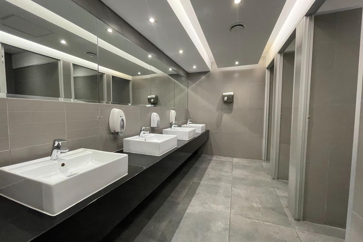 Modern commercial bathroom featuring multiple vessel sinks, a large vanity mirror, recessed lighting, and tiled finishes with partitioned toilet cubicles.