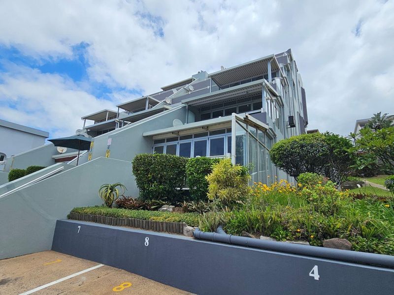Exterior view of a multi-story apartment complex featuring tiered balconies, landscaped garden beds, and designated parking bays.