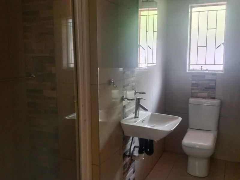 A functional bathroom featuring a shower enclosure, wall-mounted basin, and toilet with tiled finishes and a window with security bars.