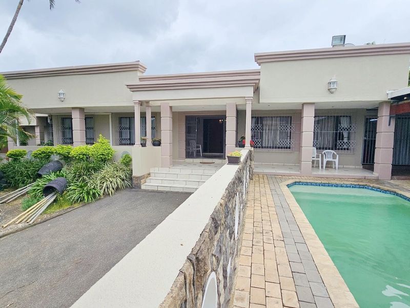 Rear exterior view featuring a swimming pool, paved deck, and a covered patio area with stone-clad retaining wall.