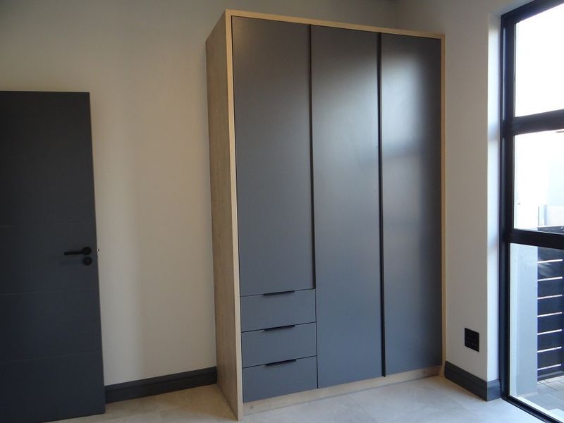 A modern bedroom featuring a sleek built-in wardrobe with integrated drawers, neutral tiled flooring, and a large black-framed window.