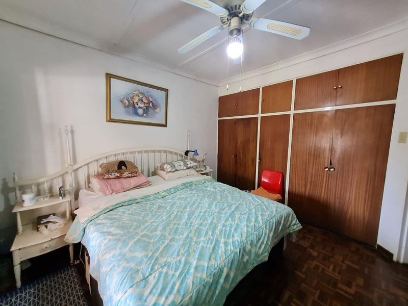 A bedroom featuring parquet flooring, built-in wooden cupboards, and a ceiling fan, offering a functional space with potential for modern updates.