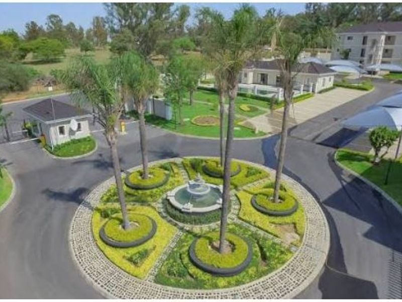 Aerial view of the townhouse complex entrance, featuring a landscaped roundabout with a central fountain, security gatehouse, and covered parking bays.