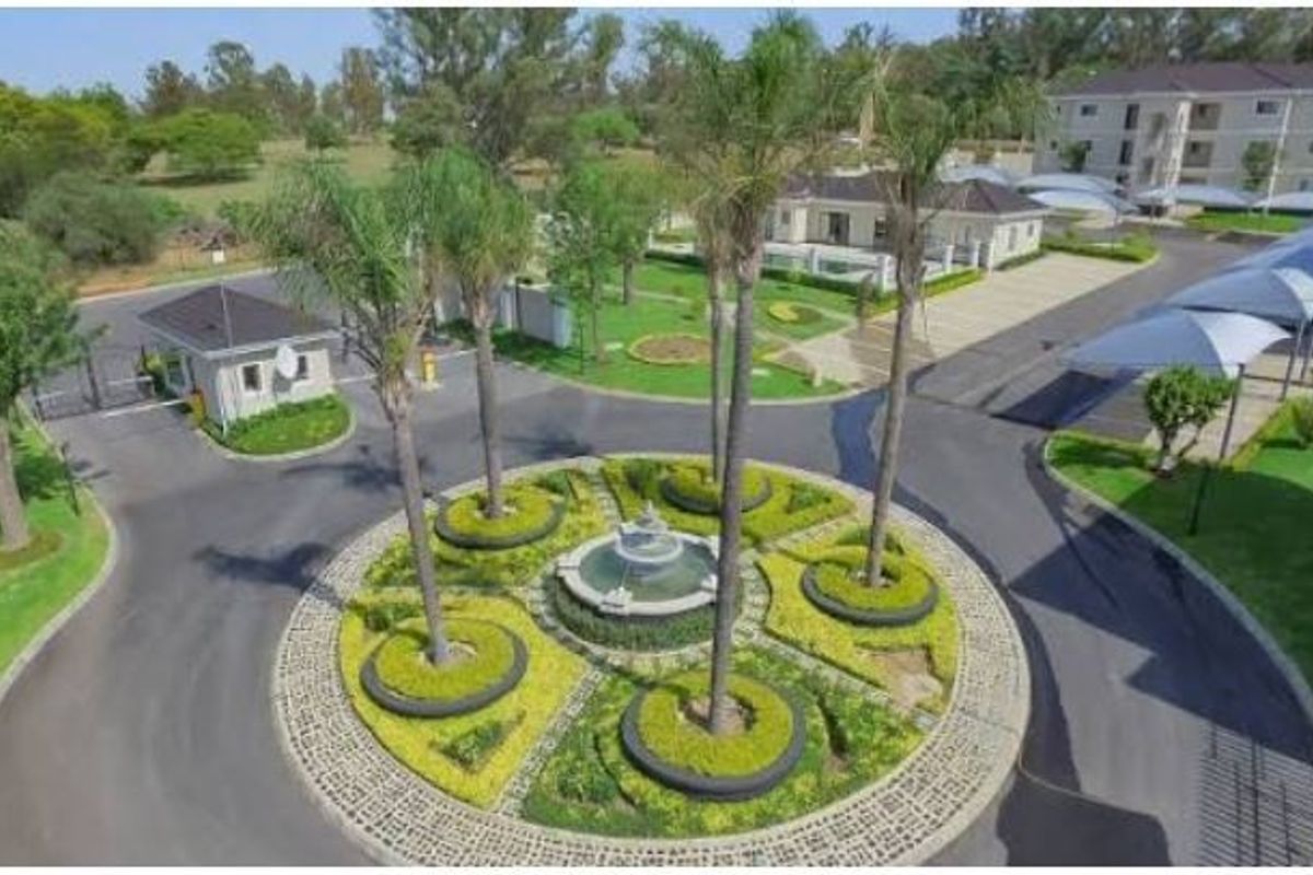Aerial view of the townhouse complex entrance, featuring a landscaped roundabout with a central fountain, security gatehouse, and covered parking bays.