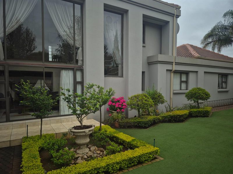 A well-maintained rear exterior view featuring a landscaped garden with artificial lawn, trimmed hedges, and a tiled patio leading to large glass sliding doors.
