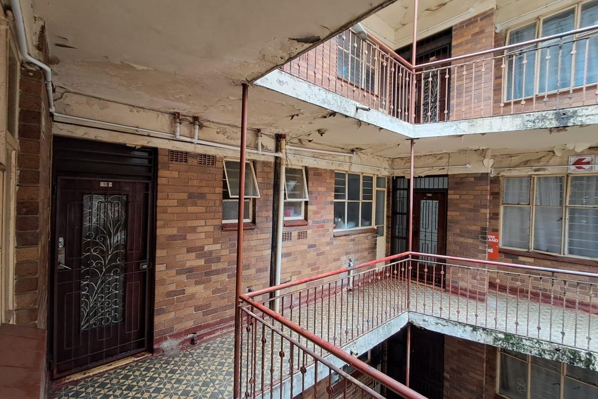 Exterior view of an apartment building walkway featuring brick facade, metal security gates, and tiled flooring, showing significant signs of wear and maintenance requirements.