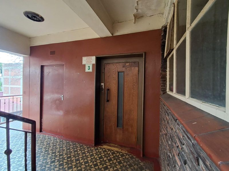A communal apartment hallway featuring a wooden entrance door, patterned floor tiles, and visible signs of wear on the ceiling and wall finishes.