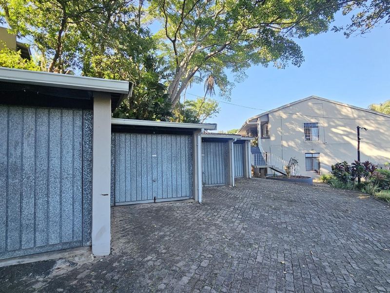 Exterior view of a row of metal garage units with paved driveway and surrounding mature trees in a townhouse complex setting.