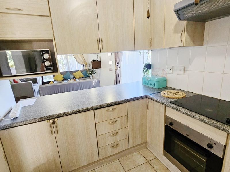 Compact kitchen featuring light wood-grain cabinetry, granite-look countertops, built-in oven, electric hob, and extractor fan, with an open-plan layout overlooking the living area.