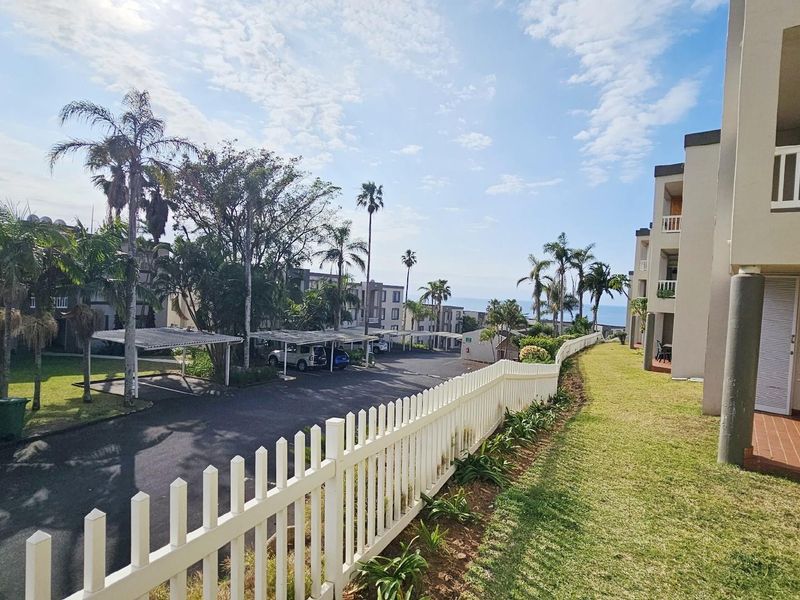 View of the apartment complex grounds featuring paved parking areas, covered carports, landscaped lawns, and a white picket fence.