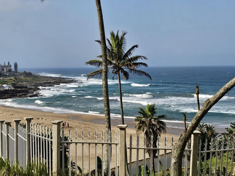 A scenic coastal view overlooking the sandy beach and ocean, framed by palm trees and a secure perimeter fence.