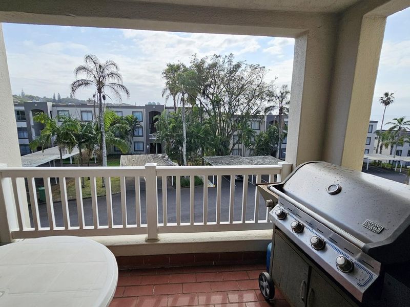 A covered balcony featuring a built-in brick braai area, white railing, and tiled flooring, overlooking the apartment complex grounds.