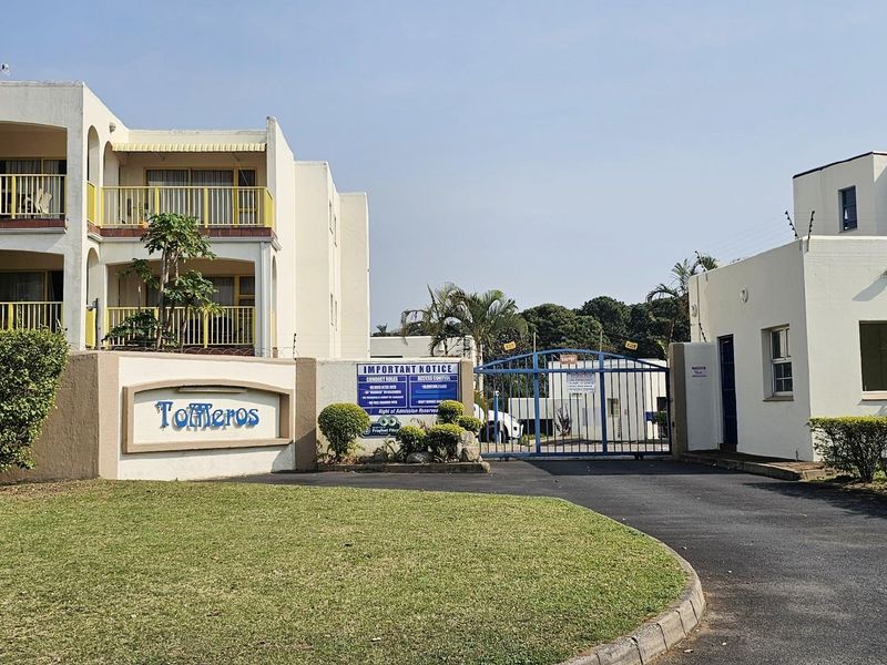 Front entrance view of the Tolmeros apartment complex, featuring a gated security access point, paved driveway, and multi-story residential buildings with balconies.