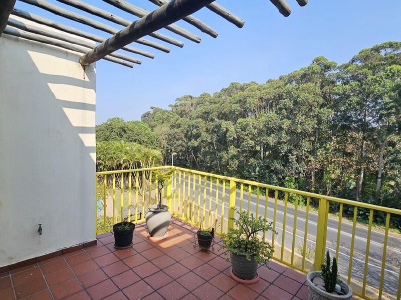 A private balcony featuring terracotta floor tiles, a timber pergola overhead, and a yellow metal safety railing overlooking a lush green treeline.