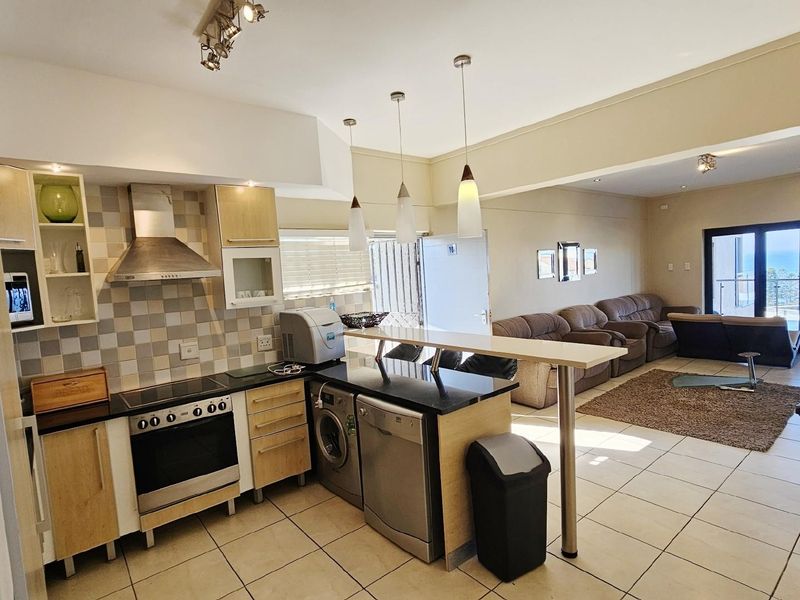 Open-plan kitchen and living area featuring a breakfast bar, built-in oven, hob, extractor fan, and tiled flooring, leading to a balcony with ocean views.