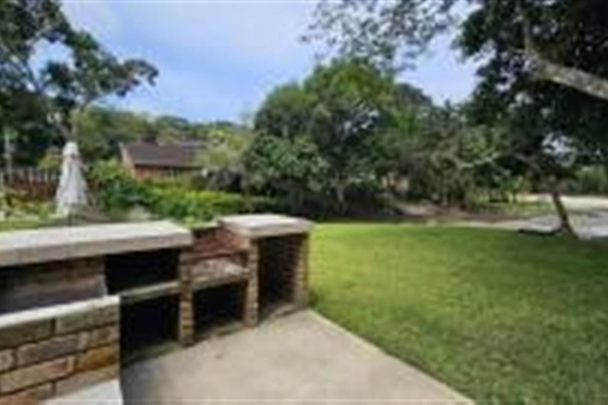 A private backyard featuring a built-in brick braai area, concrete patio, and a spacious lawn surrounded by mature trees.
