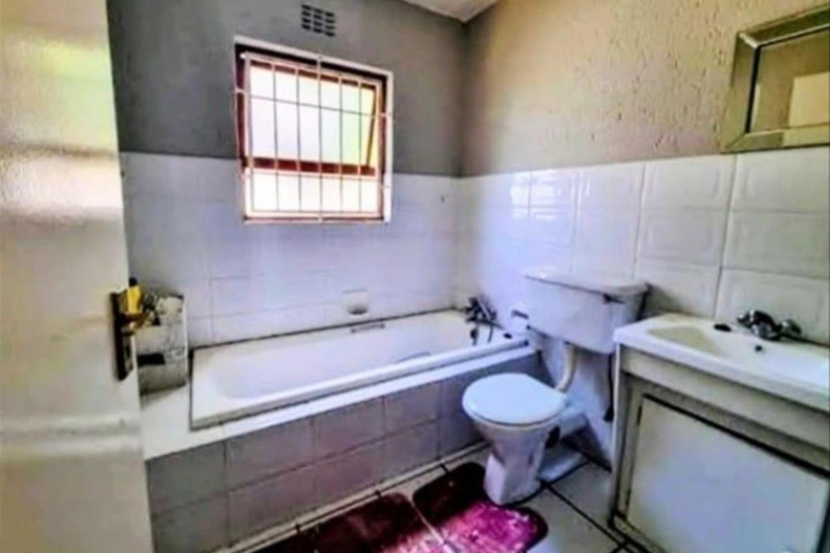 A compact bathroom featuring a built-in bathtub, a standard toilet, and a vanity basin with tiled walls and security bars on the window.