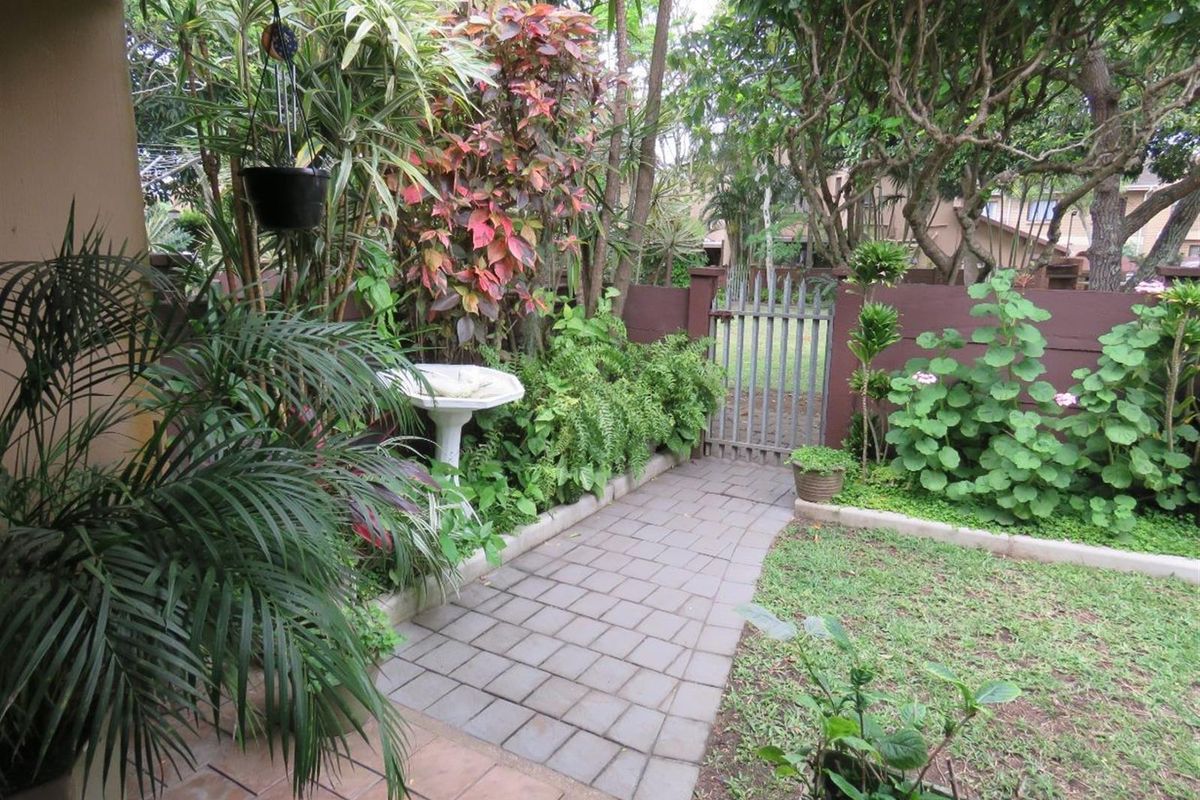 A private rear garden featuring a paved walkway, established landscaping, and a secure metal gate providing access to the exterior area.
