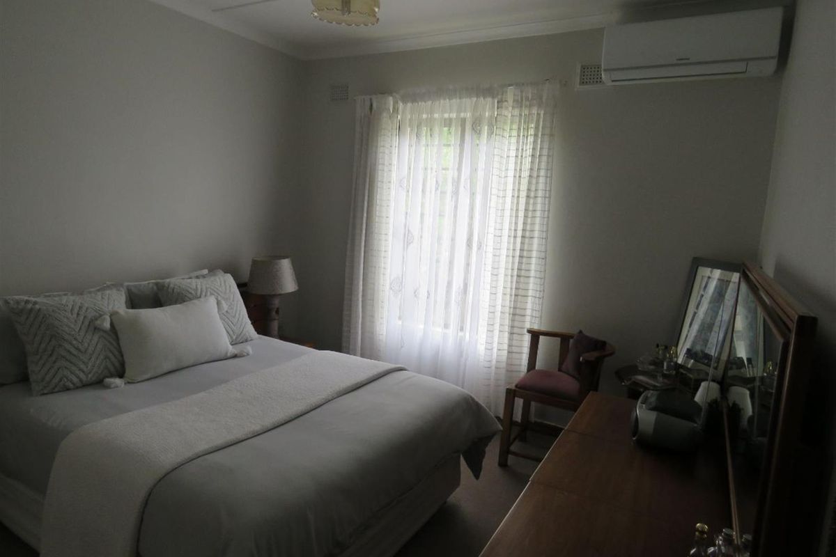 A bedroom featuring a wall-mounted air conditioning unit, neutral wall finishes, and a window with sheer curtains.
