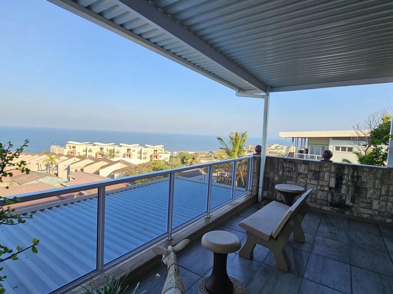 A spacious covered balcony featuring tiled flooring, a glass balustrade, and stone-clad feature wall, offering expansive coastal and neighbourhood views.