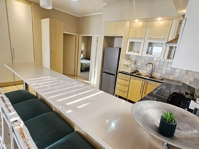 Open-plan kitchen featuring a long breakfast bar, light wood cabinetry, integrated sink, and tiled backsplash, flowing into the adjacent living and bedroom areas.