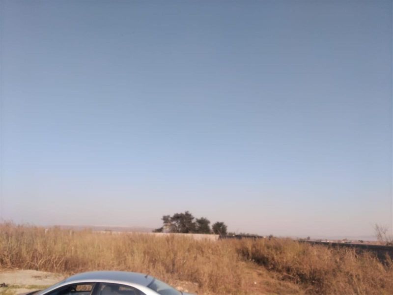 A view of a vacant residential plot featuring dry grass and a boundary wall in the distance under a clear sky.