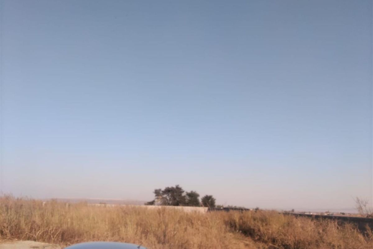 A view of a vacant residential plot featuring dry grass and a boundary wall in the distance under a clear sky.