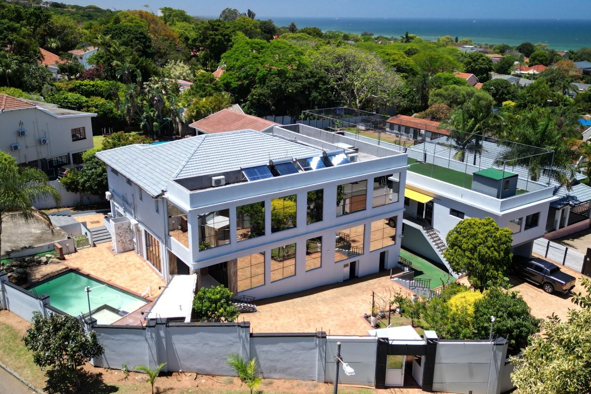 Aerial view of a modern multi-story property featuring a swimming pool, paved driveway, solar panels, and extensive perimeter walling in a coastal setting.