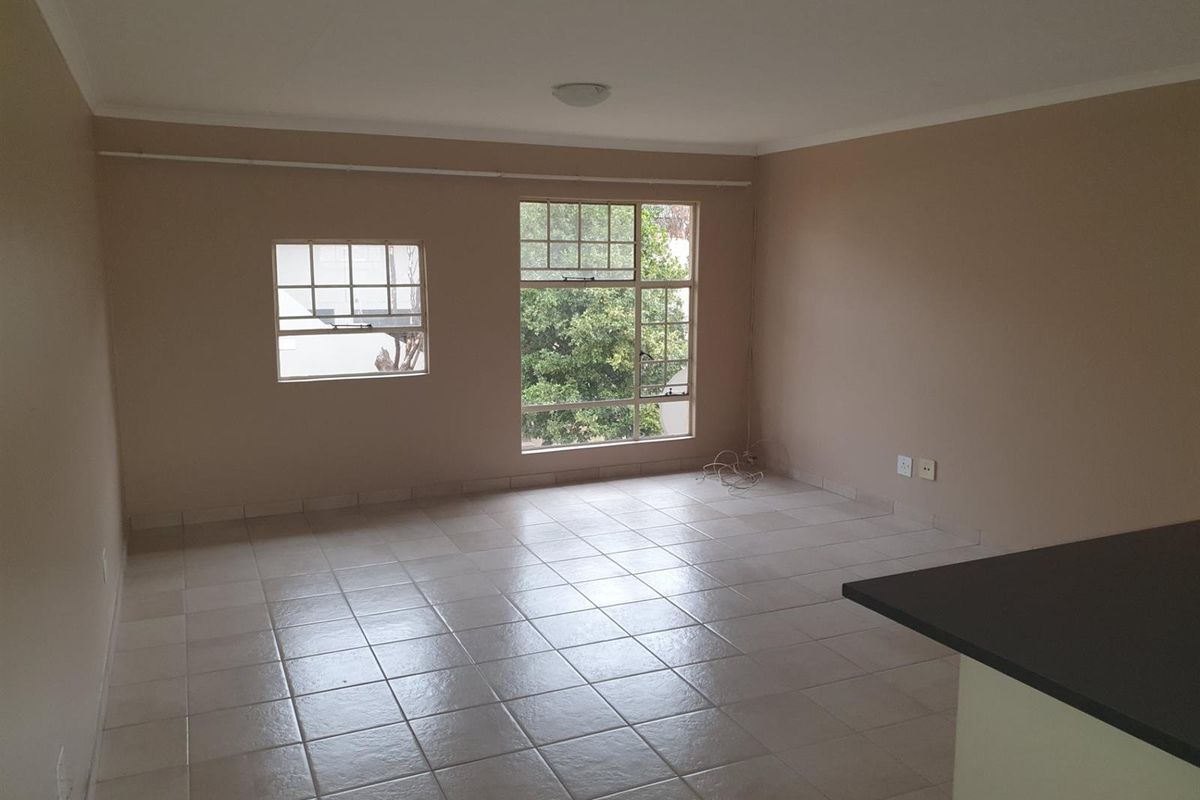 An empty, open-plan living area featuring neutral wall tones, tiled flooring, and large windows providing natural light.
