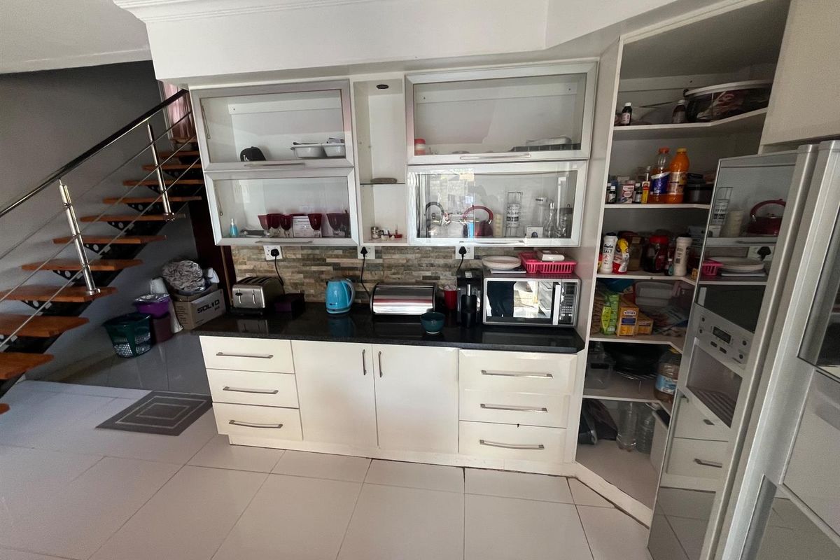 A functional kitchen area featuring white cabinetry, stone countertops, a tiled backsplash, and an integrated pantry, adjacent to an open staircase.