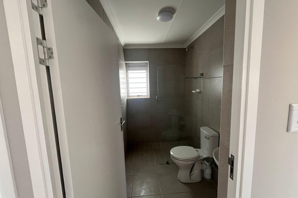 A compact bathroom featuring neutral floor-to-ceiling wall tiles, a walk-in shower enclosure, a standard toilet, and a small basin.