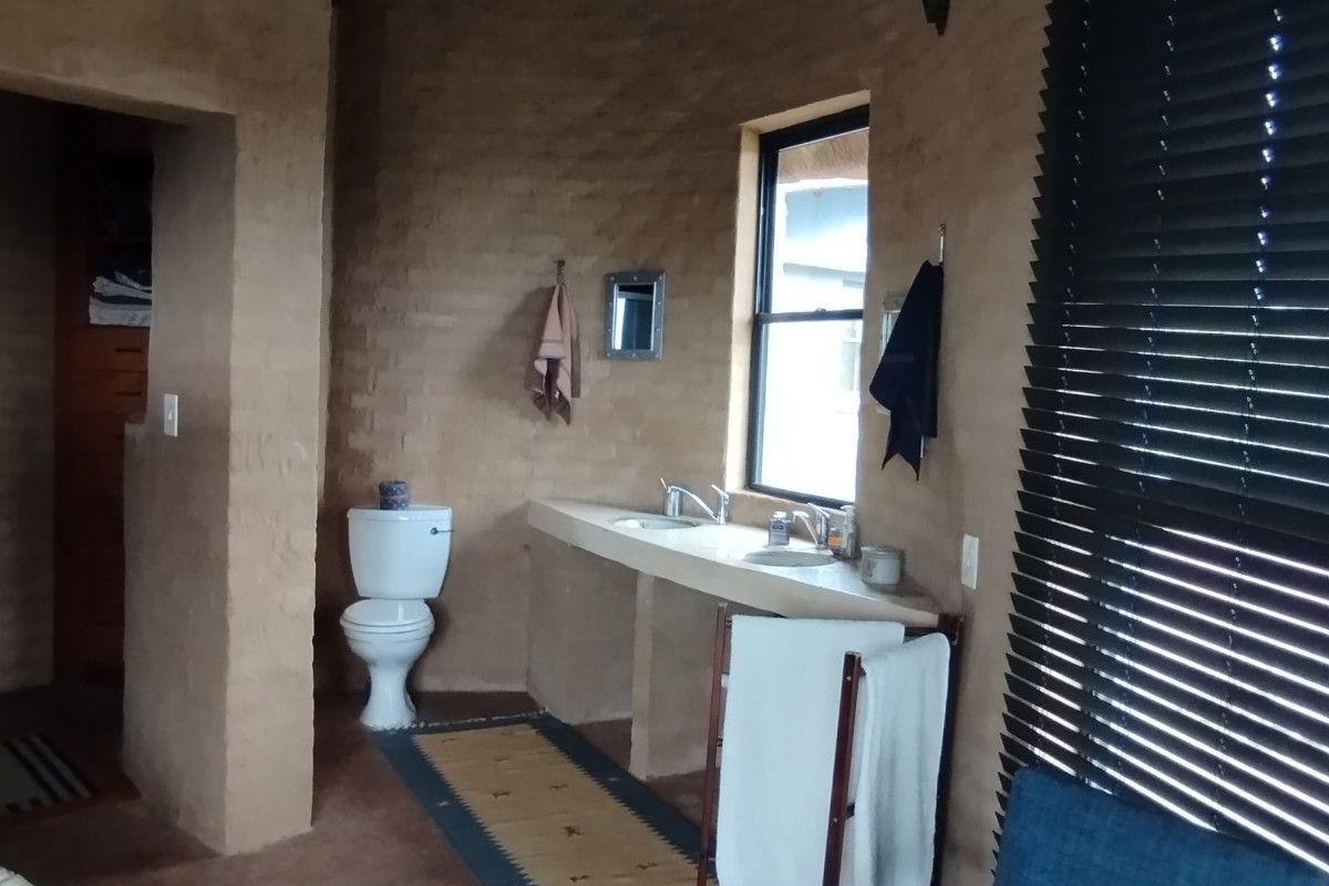 Rustic bathroom featuring a dual-basin concrete vanity, exposed thatched roof elements, and textured earth-toned walls with a private toilet area.
