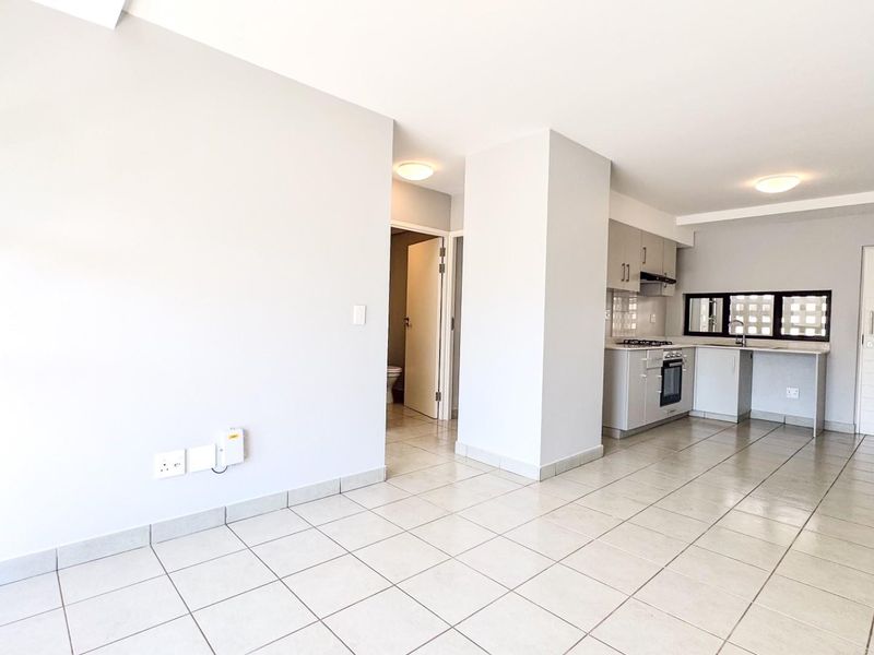An open-plan living and kitchen area featuring neutral tiled flooring, modern cabinetry, and integrated kitchen appliances in a clean, well-maintained space.