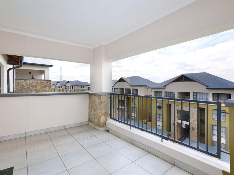 A spacious, covered balcony featuring tiled flooring, stone cladding accents, and a metal balustrade, offering views of the surrounding apartment complex.