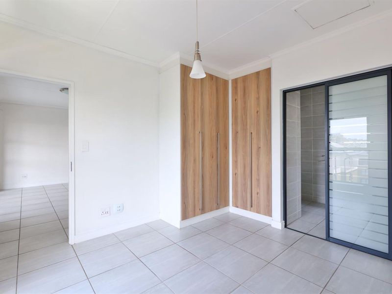 A bright, empty bedroom featuring modern wood-finish built-in cupboards, neutral tiled flooring, and a sliding glass door leading to an adjacent space.