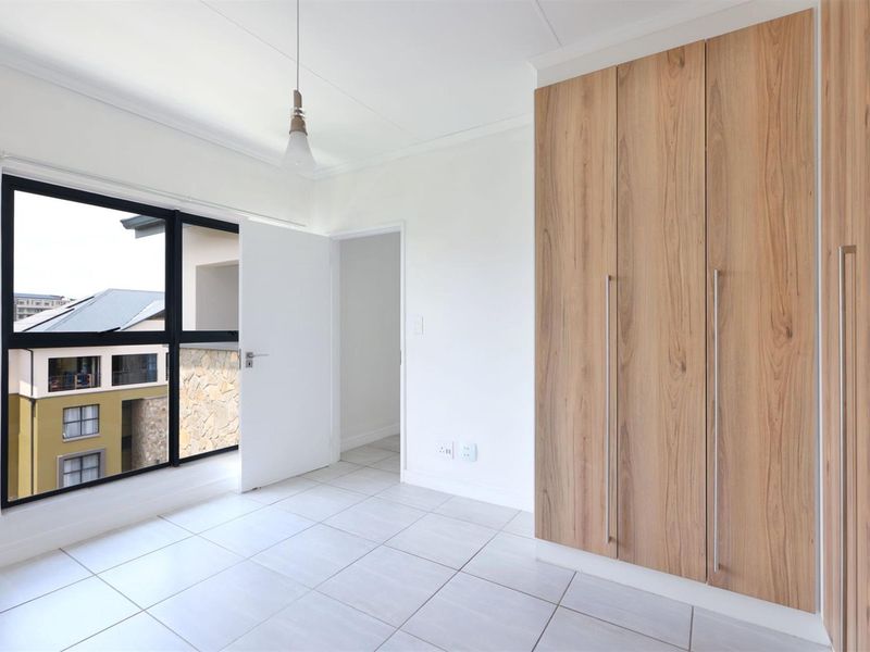 A bright, empty bedroom featuring modern wood-finish built-in cupboards, neutral tiled flooring, and a large window with views of the surrounding complex.