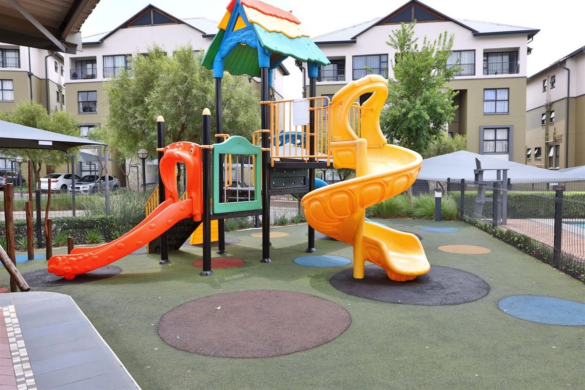 A well-maintained communal playground area featuring modern play equipment, rubberized safety surfacing, and perimeter fencing within an apartment complex.